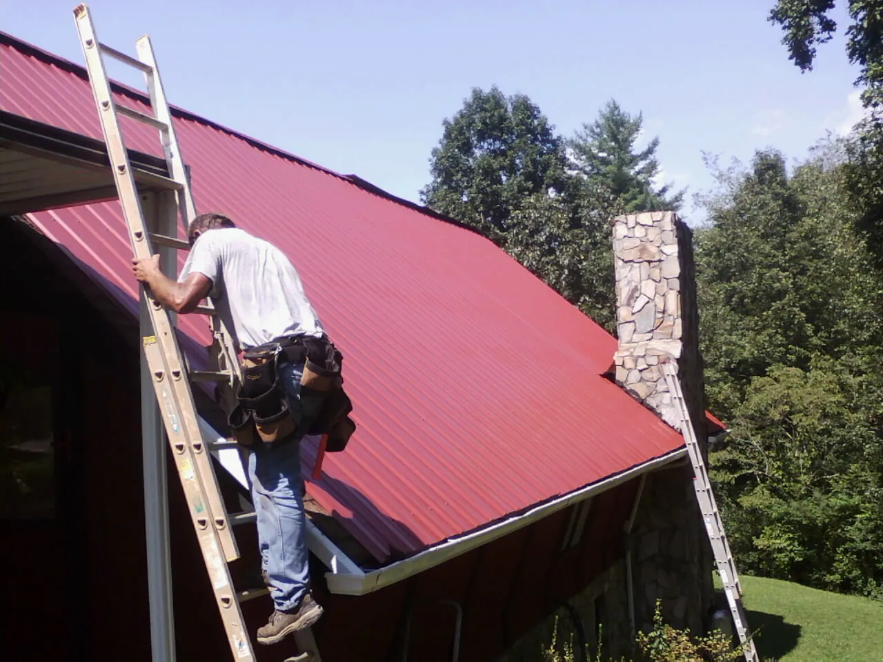 Roofer on a ladder beside a red metal roof and stone chimney — Soffit & Fascia Repair by Roofing Brothers in Saddle Brook